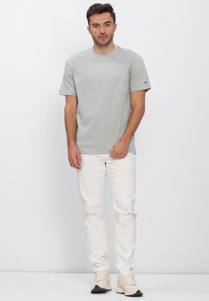 Mand står mod hvid baggrund iført lysegrå T-shirt, hvide bukser og beige sneakers, med hænder afslappet ned langs siden.