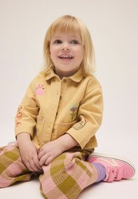 Uśmiechnięte młode dziecko o blond włosach siedzące po turecku, ubrane w żółtą kurtkę z haftem oraz w kratkowane spodnie i różowe tenisówki.