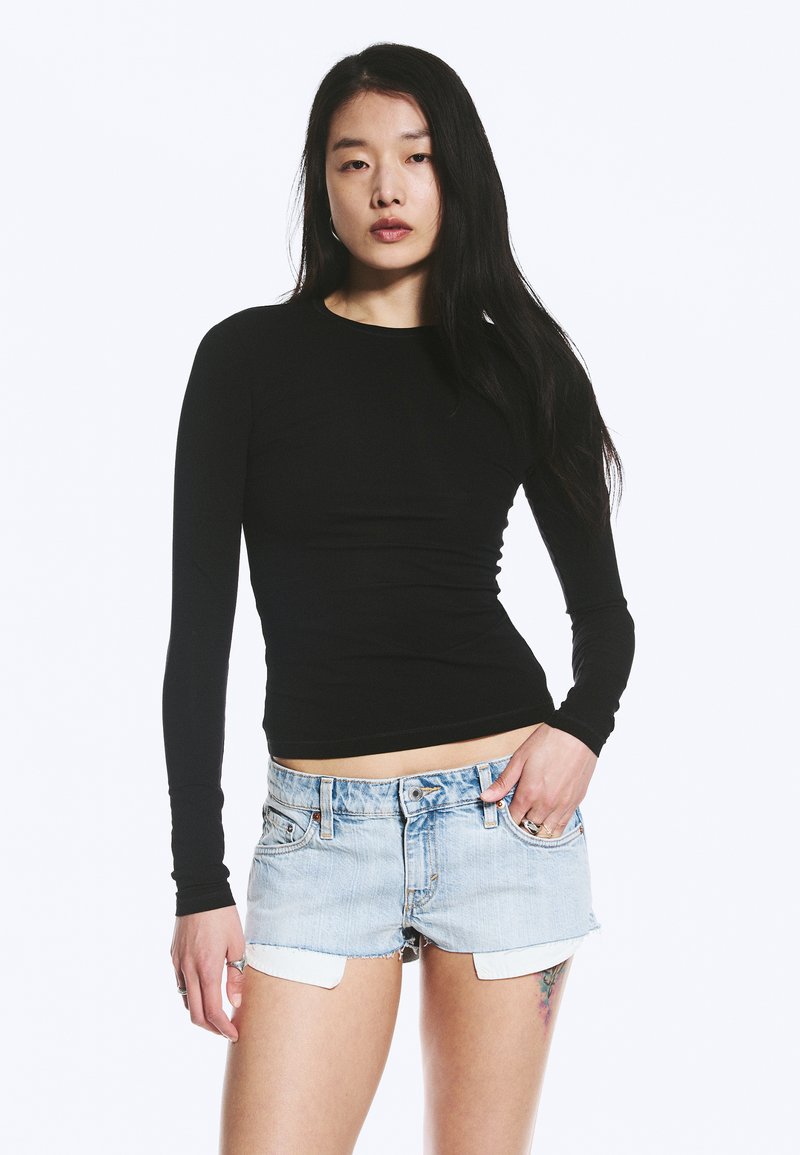 Jeune femme aux longs cheveux noirs portant un haut noir ajusté à manches longues et un short en jean bleu clair, debout avec une main dans la poche.