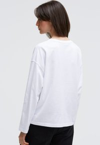 Weißes langärmliges T-Shirt mit rundem Halsausschnitt, lockerer Passform und leicht abgesenkten Schultern. Glatter Baumwollstoff, keine sichtbaren Muster.