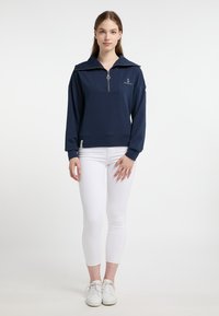 Donkerblauwe zip-up sweatshirt met een hoge kraag, voorzien van een voorzak en een ankerlogo. Gecombineerd met witte skinny jeans en witte sneakers.