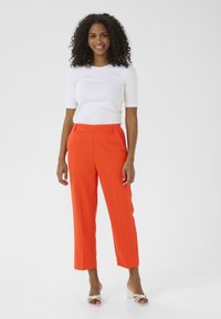 Pantaloni arancioni su misura con vita alta e gamba dritta, abbinati a una blusa bianca aderente a maniche corte. Tessuto liscio, senza motivi visibili.