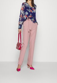Blouse longue à manches longues à fleurs bleu marine avec des couleurs variées, associée à un pantalon tailleur rose clair et des chaussures à talons roses, tenant un petit sac à main.