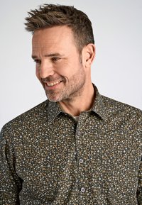 Camisa de botones con un fondo oscuro y un patrón floral en tonos blancos y marrones; tejido texturizado con un cuello clásico.