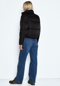 En blond person som bär en svart puffjacka, blå jeans och gröna sneakers, står vänd bort mot en vit bakgrund.