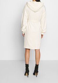 Ropa de baño con capucha en crema y cinturón de lazo, de manga larga y corte relajado, con una textura suave y largo hasta la rodilla. Botas de tacón alto en negro.