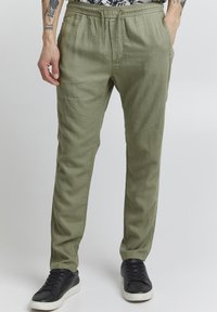 Pantalons en lin vert olive clair avec une taille élastique à cordon de serrage, coupe fuselée et poches latérales, associés à des baskets noires.