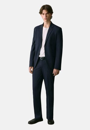 Traje marino que presenta una chaqueta estructurada con solapas de muesca y pantalones rectos. Usado sobre una camiseta blanca sencilla, combinado con zapatos negros.