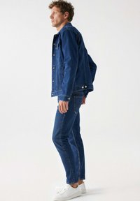 Chaqueta de mezclilla y jeans en azul oscuro, con botones frontales, bolsillos laterales y un diseño ajustado. Combinados con zapatillas blancas.
