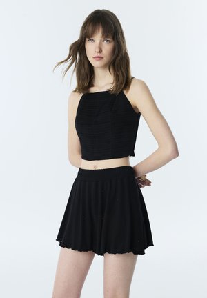 Junge Frau mit schulterlangem braunem Haar, die ein schwarzes ärmelloses Croptop und schwarze Shorts mit hoher Taille vor hellem Hintergrund trägt.