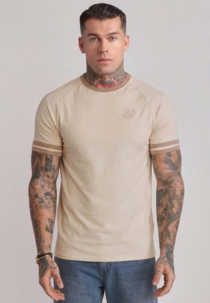 Mann mit Hals- und Arm-Tattoos, der ein beiges kurzärmeliges T-Shirt mit gestreiften Bündchen und blaue Jeans trägt, steht vor einem einfarbigen Hintergrund.