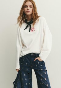 Jonge vrouw met lang rood haar, gekleed in een witte trui met rode tekst, donkerblauwe spijkerbroek met metalen ringen en een zwarte nekband.