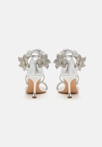 Sandales à talons hauts argentées avec des brides transparentes et des embellissements floraux en bijoux sur le talon. Caractérisées par un design élégant et une texture brillante.