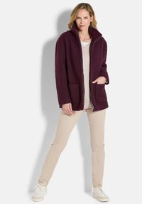 Burgunderfarbene Fleecejacke mit hohem Kragen, Reißverschluss, zwei aufgesetzten Taschen, strukturiertem Obermaterial, getragen über einem hellen Oberteil und cremefarbenen Hosen.
