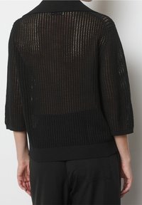 Schwarze gestrickte Strickjacke mit einem Netzmuster, Dreiviertelärmeln und einem gerippten Saum. Der Rücken ist offen für zusätzliche Atmungsaktivität.