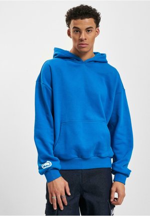 Sudadera con capucha de color azul brillante, hecha de una mezcla de algodón. Presenta un bolsillo tipo canguro y un logo blanco en la manga izquierda.