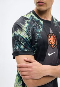 Κοντινό πλάνο ατόμου που φοράει μαύρη και πράσινη φανέλα ποδοσφαίρου KNVB με πορτοκαλί έμβλημα λιονταριού και λογότυπο Nike, με σταυρωμένα τα χέρια στο στήθος.