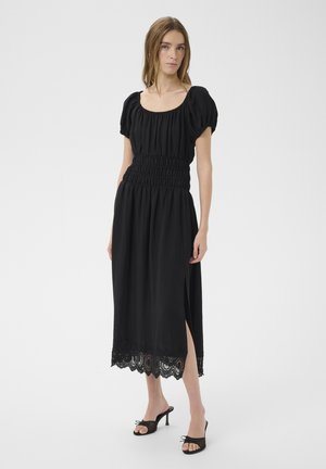 Femme portant une robe midi noire avec des manches bouffantes, une taille smockée, une fente latérale et un ourlet en dentelle, assortie à des sandales noires à talons.