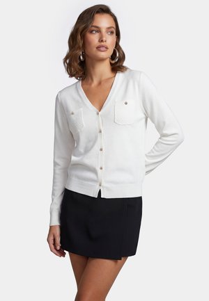 Kvinde med bølget hår iført hvid knapfast cardigan med to forlommer og et sort miniskørt, stående mod en lys baggrund.