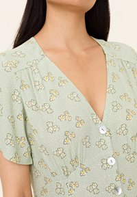 Grünes Blumenkleid mit kurzen Ärmeln, V-Ausschnitt, Knopfleiste und strukturiertem Stoff mit weißen und gelben Blumenmustern.