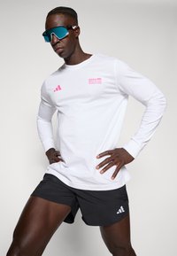 White long-sleeve shirt featuring pink BMW Berlin Marathon logo and three stripes. Paired with black shorts and blue sunglasses.