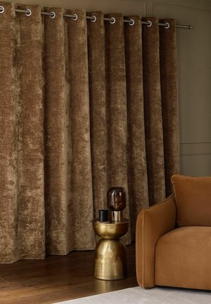 Tende di velluto marrone con occhielli argentati appese accanto a una poltrona in camoscio marrone e un tavolino dorato con oggetti decorativi.