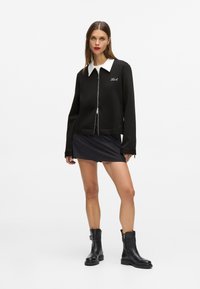Čierna zipsová bunda s bielym golierom, s logom na hrudi, skombinovaná s krátkou čiernou sukňou a čiernymi koženými členkovými topánkami.