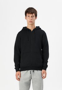 Sudadera negra con cremallera hecha de una mezcla de algodón, con capucha, bolsillos frontales, puños de canalé y un corte relajado, combinada con pantalones de chándal grises.