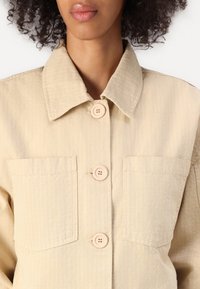 Casaco bege com colarinho, apresentando listras horizontais, dois bolsos no peito e botões grandes. O tecido parece ser texturizado e leve.