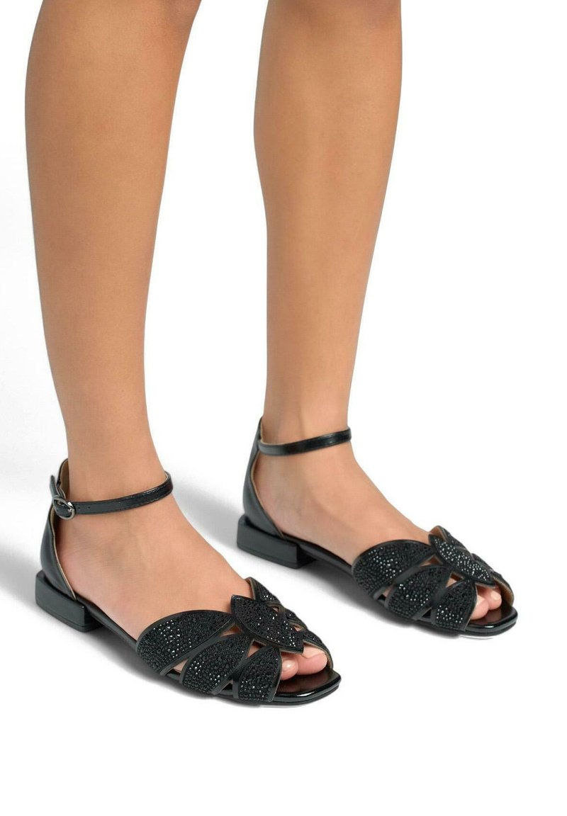 Sandales noires avec un petit talon carré, présentant des lanières croisées et un design floral agrémenté de textures. Boucle de cheville réglable incluse.