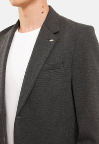 Szary żakiet o teksturowanej powierzchni z wykładanym kołnierzem, z małym emblematem ryby na lewym klapie i kieszenią na piersi. Noszony na białej koszulce.