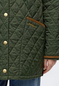 Πράσινο μπουφάν Barbour με καπιτονέ, καφέ φινίρισμα, χρυσά κουμπιά και ορατό χέρι κοντά στη μανσέτα πάνω από μπλε τζιν από denim.