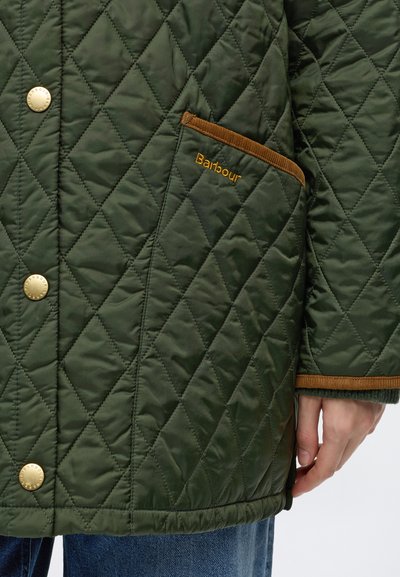Зелено капирано яке Barbour с кафяво обкантване, златни копчета и видима ръка близо до маншета върху сини дънки от деним.