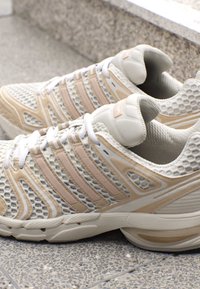 Paar beige-weiße Mesh-Laufschuhe mit weißen Schnürsenkeln, die auf einem gefliesten Boden nahe einer Steinstufe liegen.