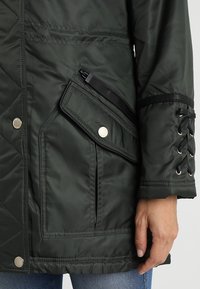 Poche de veste matelassée vert foncé avec bouton-pression et fermeture éclair, avec un détail à lacets sur la manche, portée avec un jean bleu.