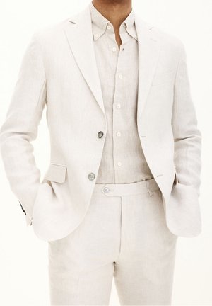 Completo in lino beige chiaro con giacca monopetto, rever a jeans e due bottoni. Abbinato a una camicia in lino e pantaloni coordinati.