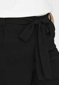 Pantalones de tiro alto negros con un acabado texturizado, que presentan un detalle de cinturón anudado en la cintura y sutiles pliegues cerca de los bolsillos.