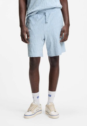 Mann trägt hellblaue, strukturierte Shorts mit elastischem Bund, weiße Socken mit blauem "HUGO"-Logo und cremeweiße Sneakers mit blauer Umrandung.