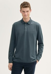 Ciemnozielona koszulka polo z długim rękawem wykonana z gładkiej tkaniny, z zapięciem na guziki przy kołnierzyku oraz subtelnym logo na klatce piersiowej.