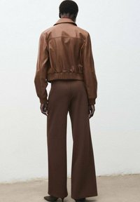 Hnědá kožená bomber bunda s ribbovanými manžetami a lemem, v kombinaci s širokými hnědými kalhotami. Hladká textura, jednoduchý design, minimální detaily.