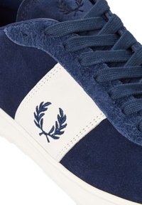 Zapatillas de ante azul marino con un parche lateral de cuero blanco que presenta un diseño de laurel bordado. Lengüeta texturada y cordones en azul marino.