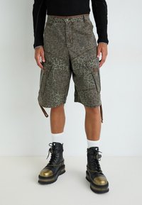 Cargo-Shorts in einem grünen Leopardenmuster mit mehreren großen Taschen, einem verstellbaren Saum und kombiniert mit schwarzen Stiefeln mit goldenen Kappen.