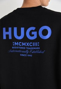 Sudadera negra con un gran texto azul que dice "HUGO", con texto gráfico azul adicional que detalla la información de la marca registrada debajo.