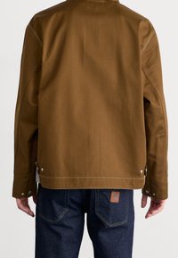 Achteraanzicht van een persoon in een bruine jas met knoopmanchetten en donkerblauwe jeans met zichtbare stiksels en een leren merklabel.