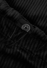 Melns korduroja audums ar vertikālām ribām, jostasvietā arapaļa melna pogu un elastīgu joslu regulējamai piemērotībai.