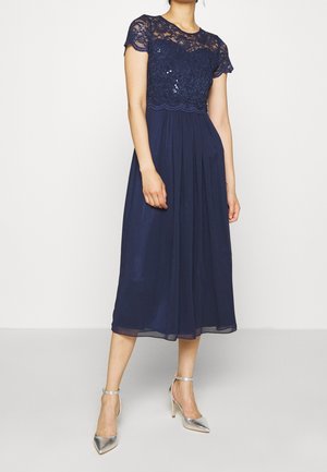 Femme portant une robe midi bleu marine avec un corsage en dentelle et paillettes et des manches courtes transparentes, associée à des chaussures à talons hauts argentées.