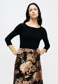 Μαύρο μακρυμάνικο τοπ με φαρδιά λαιμόκοψη σε συνδυασμό με μαύρη φούστα με χρυσό λουλουδάτο σχέδιο και μεσαίο μήκος.