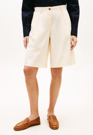 Person iført cremefarvede knælange shorts, sort langærmet skjorte og brune læder slip-on loafers, stående på hvid baggrund.