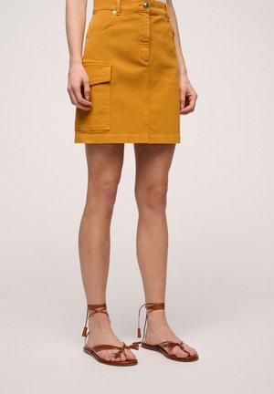 Katoenen denim minirok in mosterdgeel met voorzakken en een aansluitend ontwerp, gecombineerd met bruine sandaaltjes met bandjes en platte zolen.