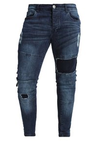 Mörkblå denimjeans med slitna detaljer, inklusive revärer vid knäna och texturerade lappar. Slimmad design med fem fickor och knappstängning.